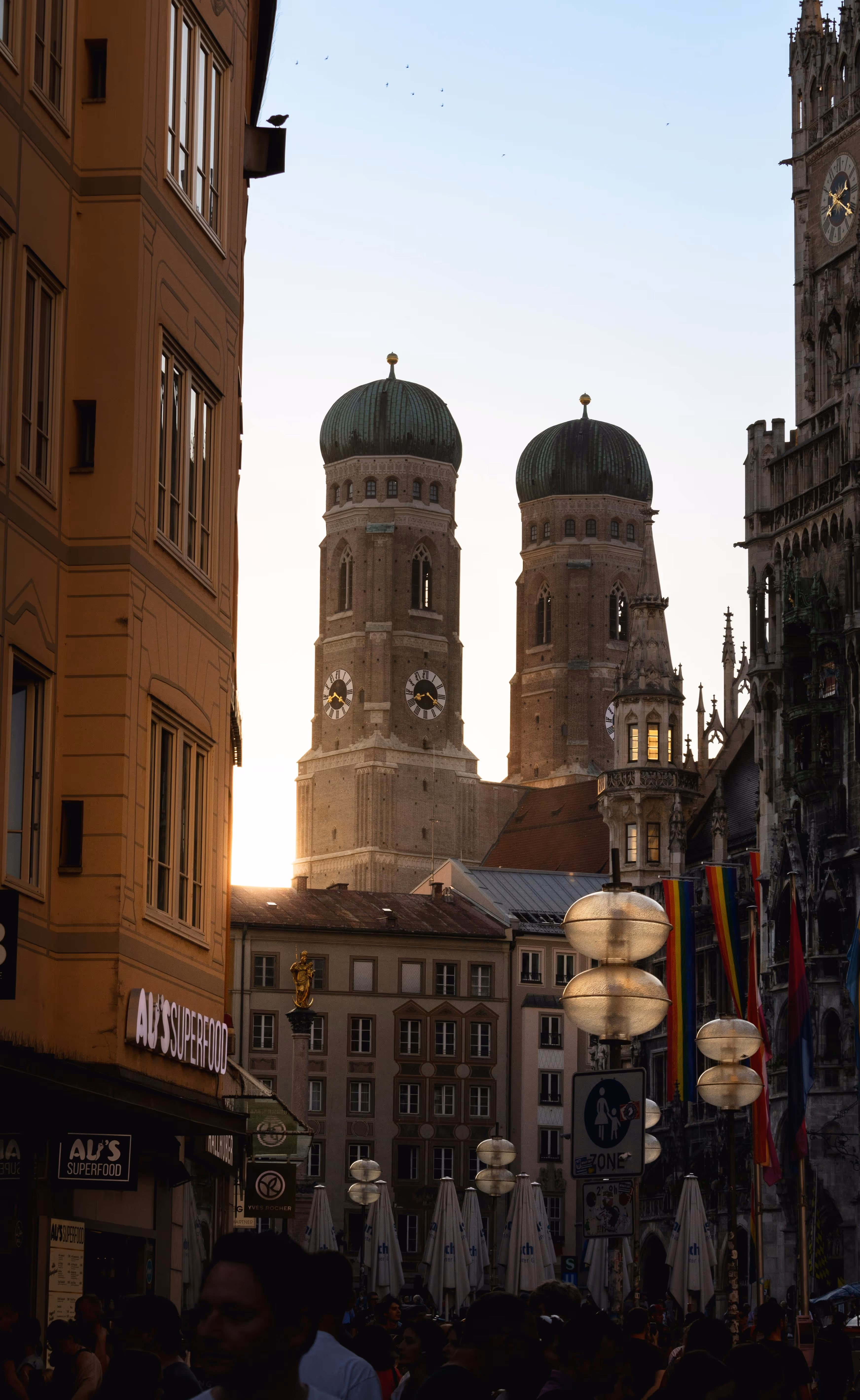 Evening view of Munich's Frauenkirche twin towers with green domes behind city buildings and a crowd on the street.
