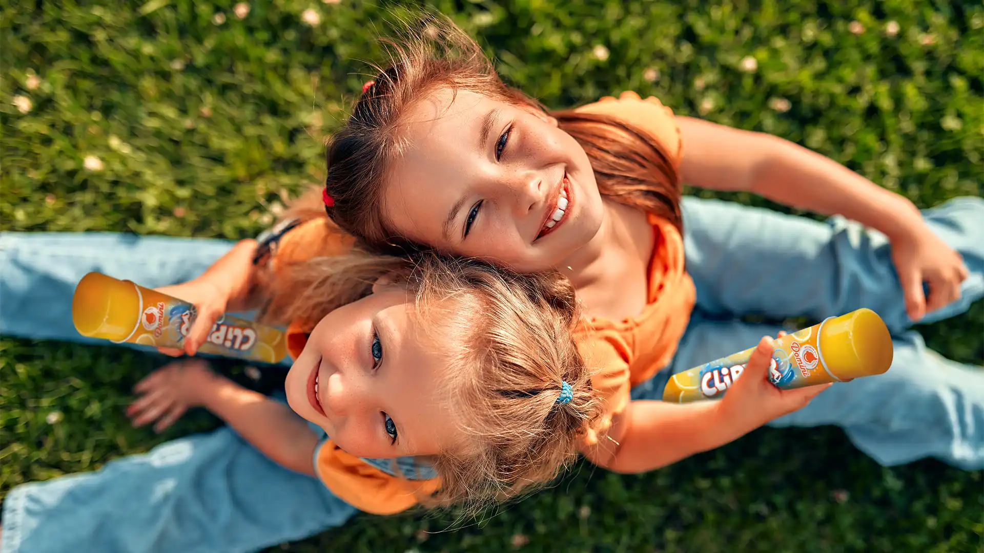 Twee lachende meisjes zittend op gras met oranje tops en blauwe jeans, elk een geel ijsje vasthoudend.