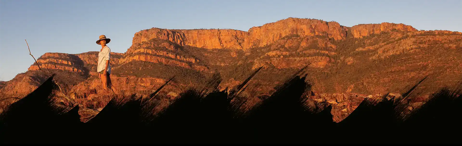 Persoon in licht shirt en hoed staat in droog grasland met op de achtergrond een rotsachtig berglandschap bij zonsondergang.