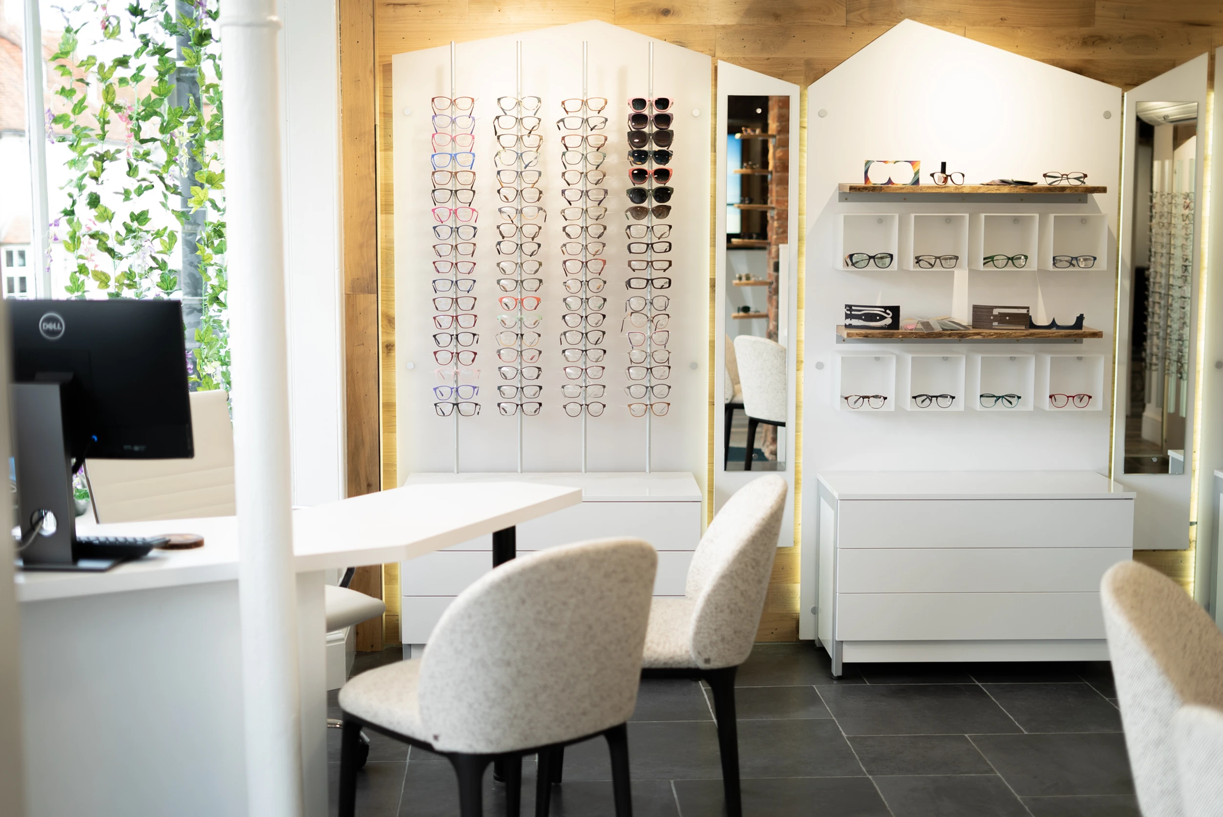 Interior of an eyeglass store with multiple eyeglass frames displayed on white stands against wooden walls and cream upholstered chairs around a white table.