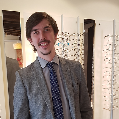 Young man in a gray suit and blue polka dot tie smiling inside an eyewear store with glasses displayed on stands behind him.