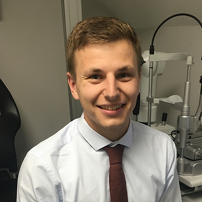 Young man with light brown hair wearing a white shirt and maroon tie, smiling in an office setting with ophthalmic equipment in the background.