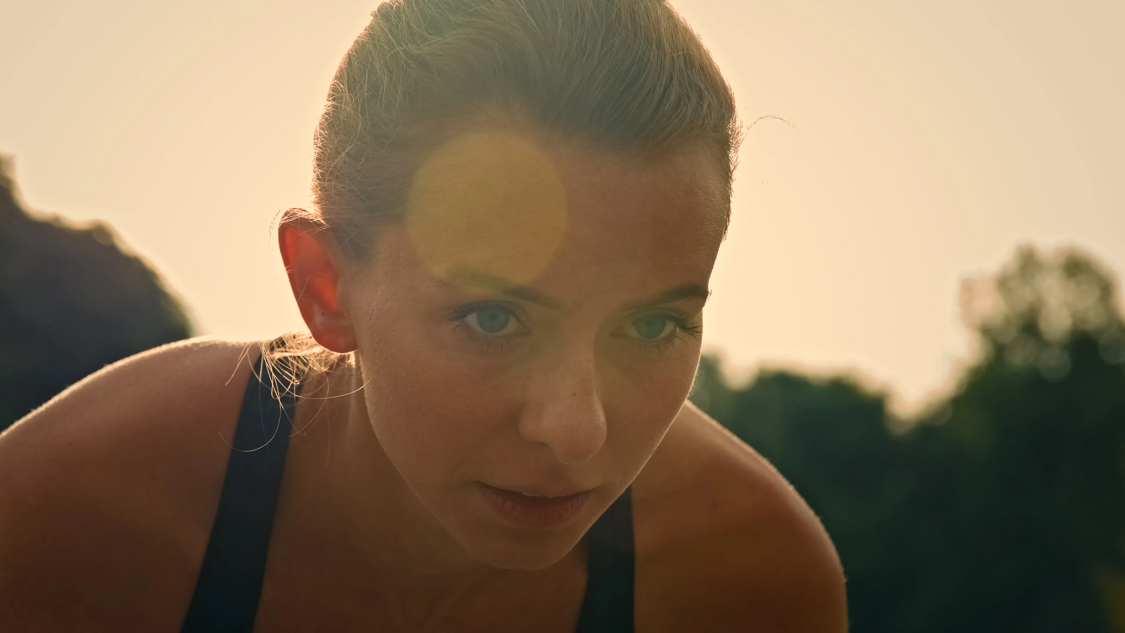 Close-up of a focused woman outdoors with sunlight and lens flare, wearing a black tank top.