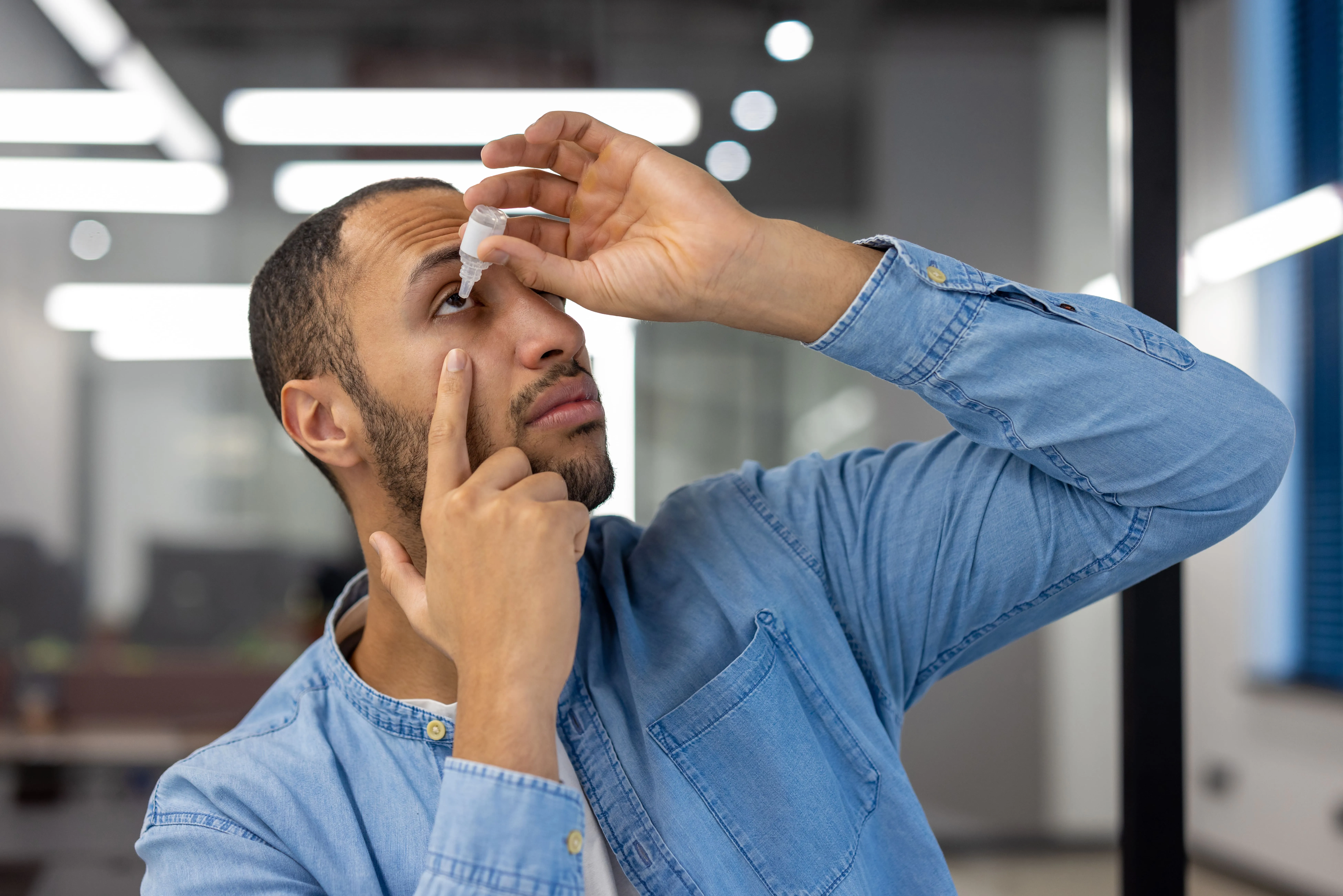 Man in a light blue shirt applying eye drops to his left eye indoors.