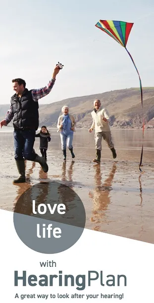 Family of four joyfully running on a beach with a colorful kite flying in the sky, promoting HearingPlan.