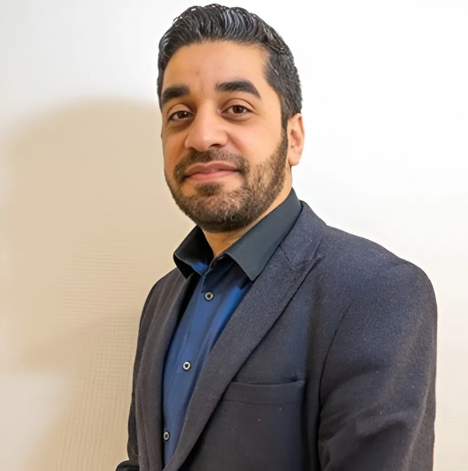 Man with neatly styled dark hair and beard wearing a dark blazer and blue shirt, smiling slightly against a plain white background.