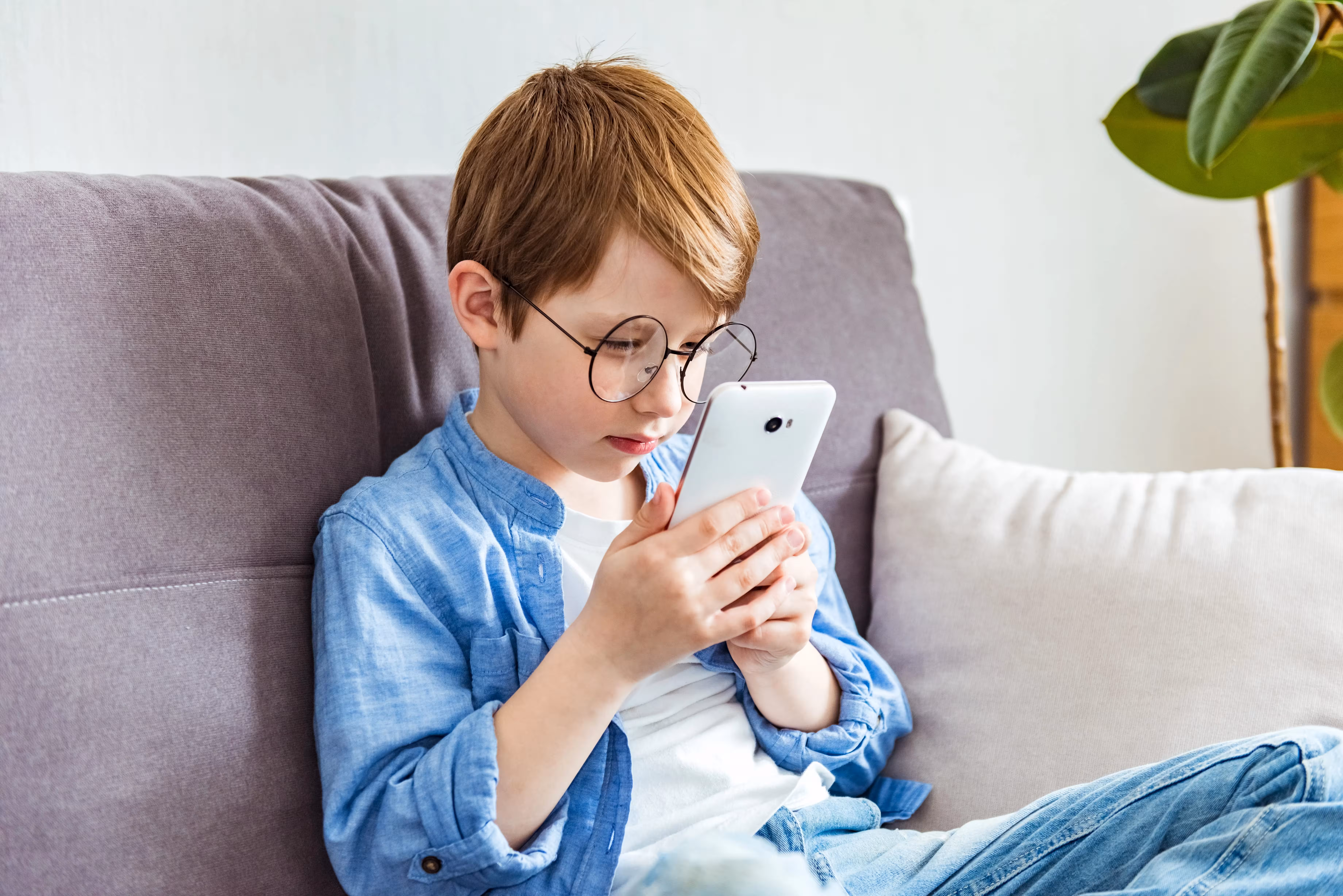 Young boy wearing glasses intently looking at a smartphone while sitting on a couch.