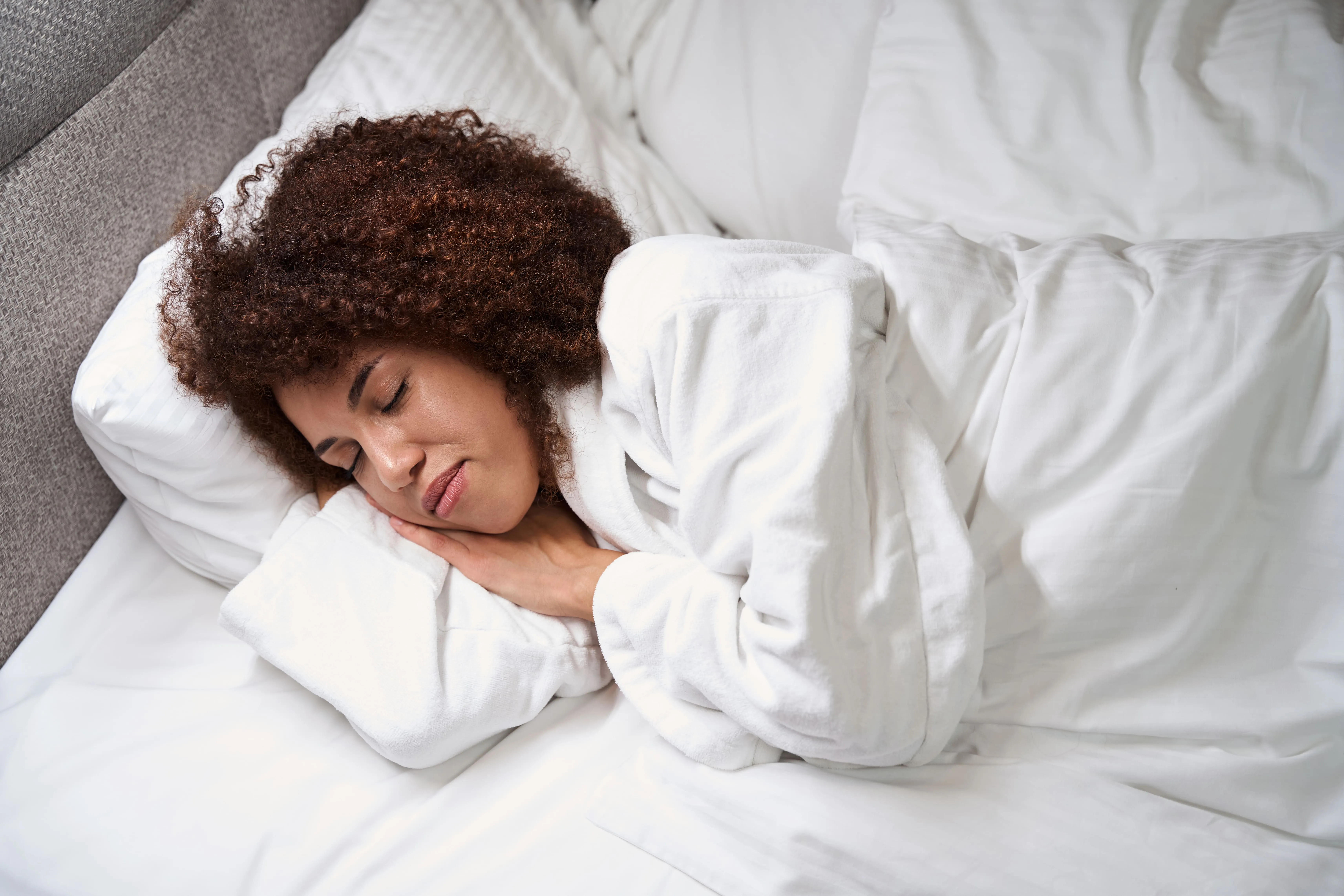 Woman with curly hair sleeping comfortably on a white bed wearing a white bathrobe.
