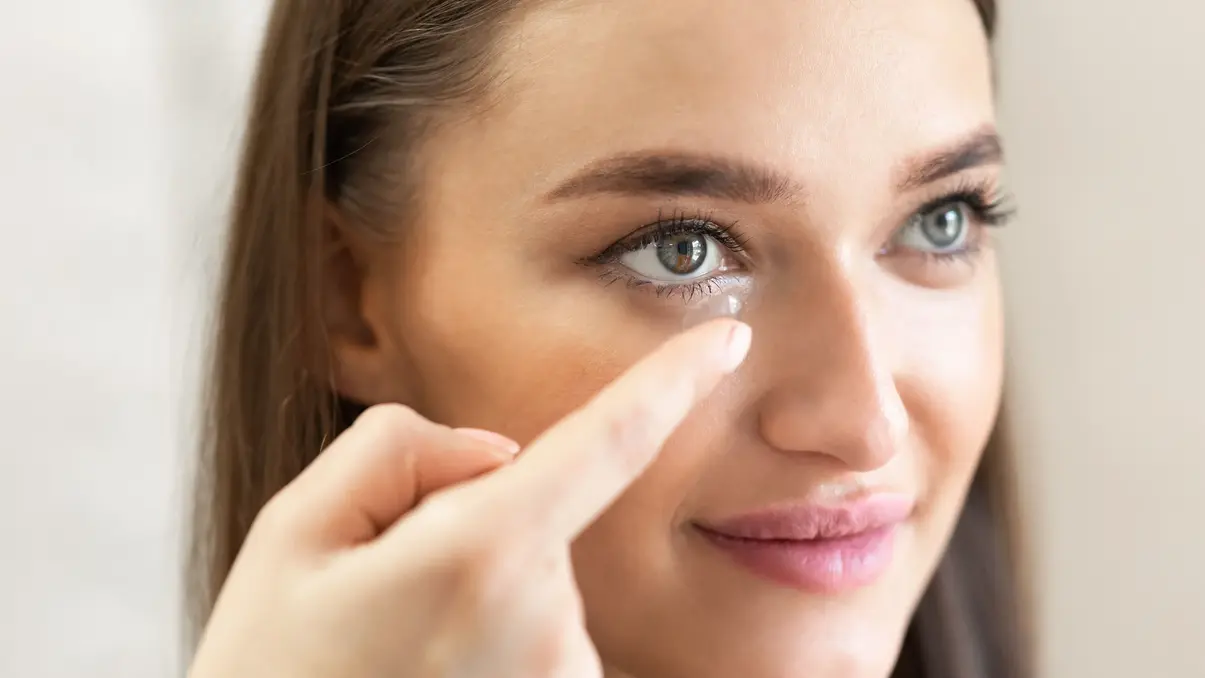 Close-up of a woman with blue eyes smiling as she places a contact lens on her finger near her eye.