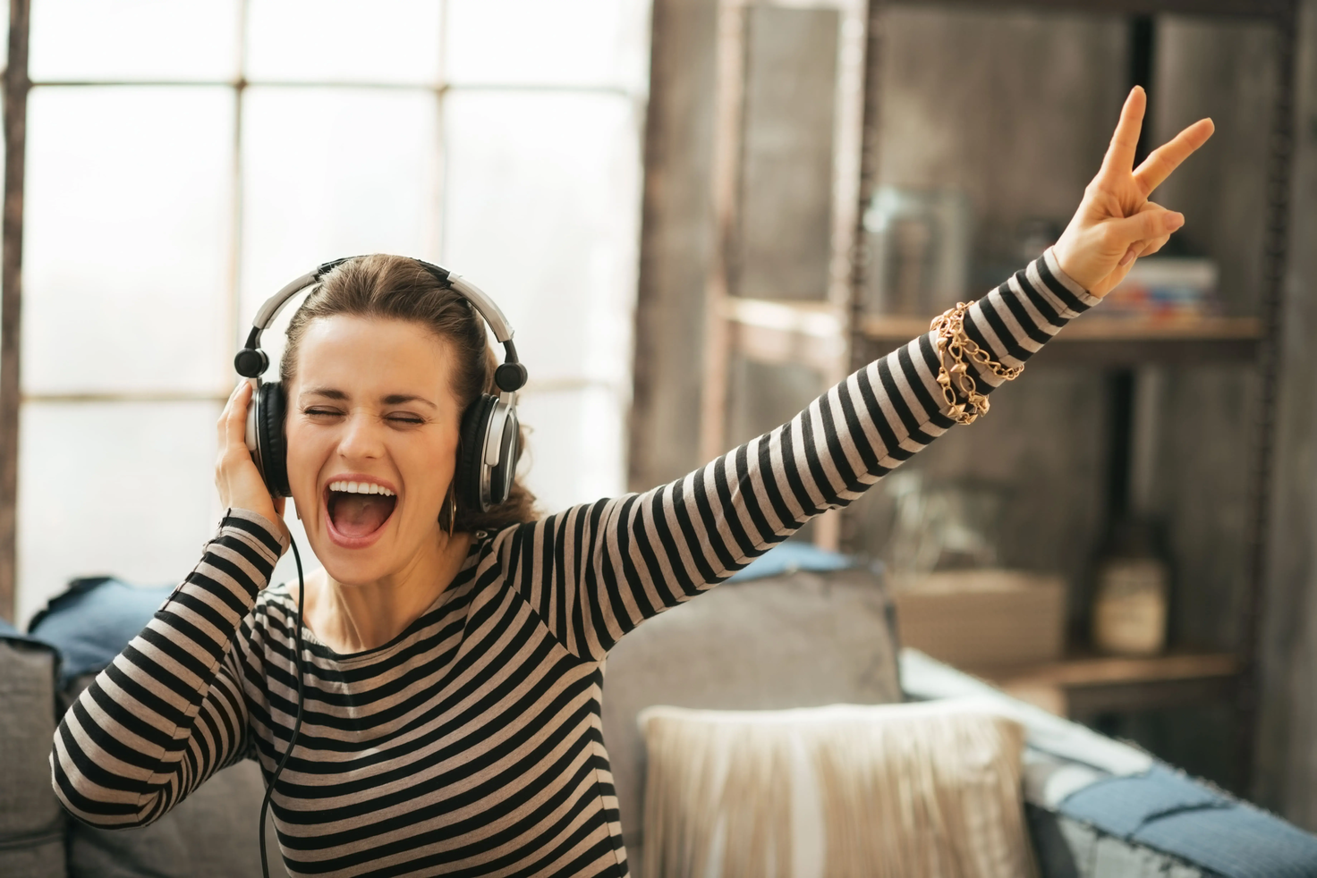 Woman wearing headphones, singing with eyes closed and making a peace sign with her hand.