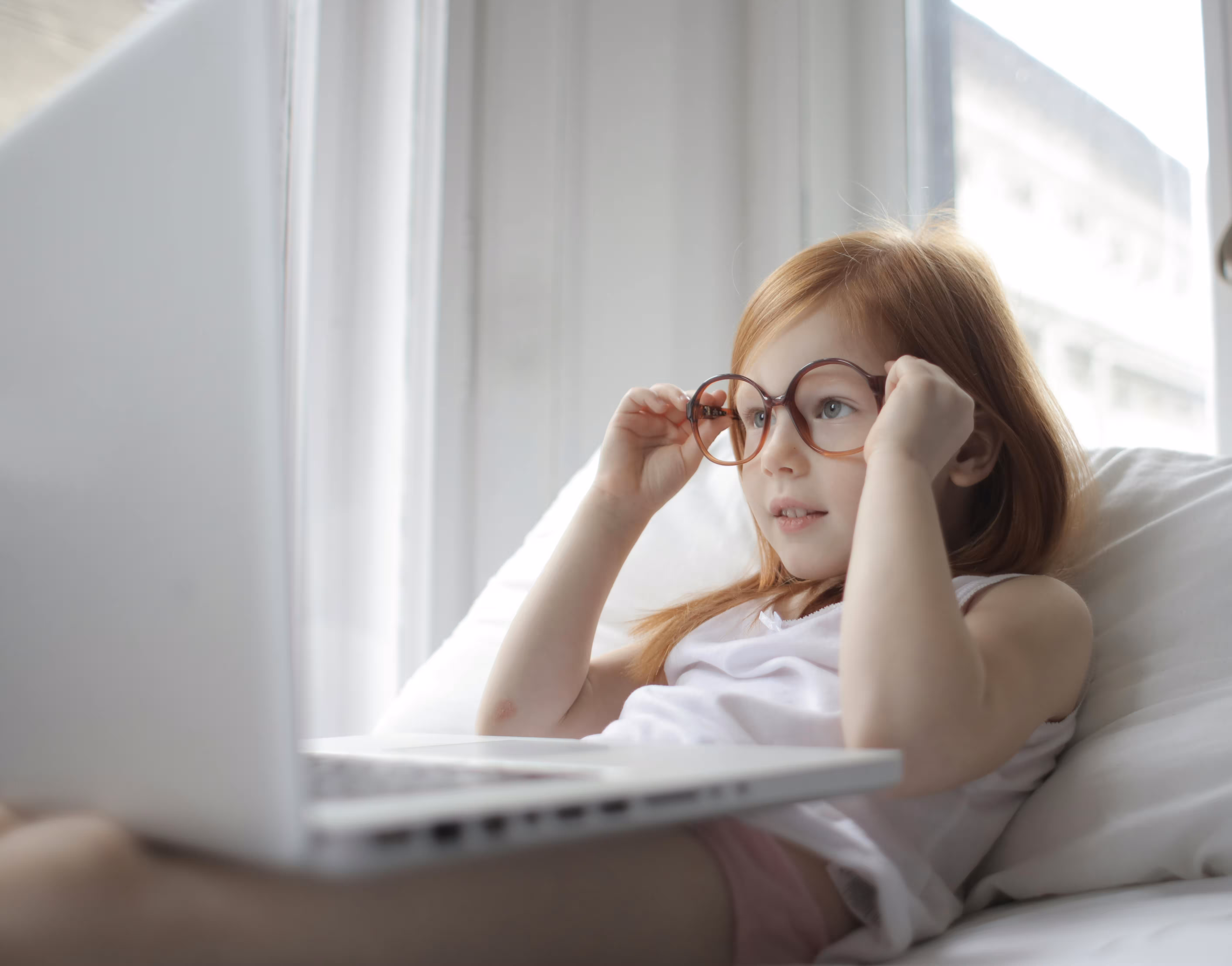 Young red-haired girl wearing large glasses using a laptop while sitting on a bed near a window.