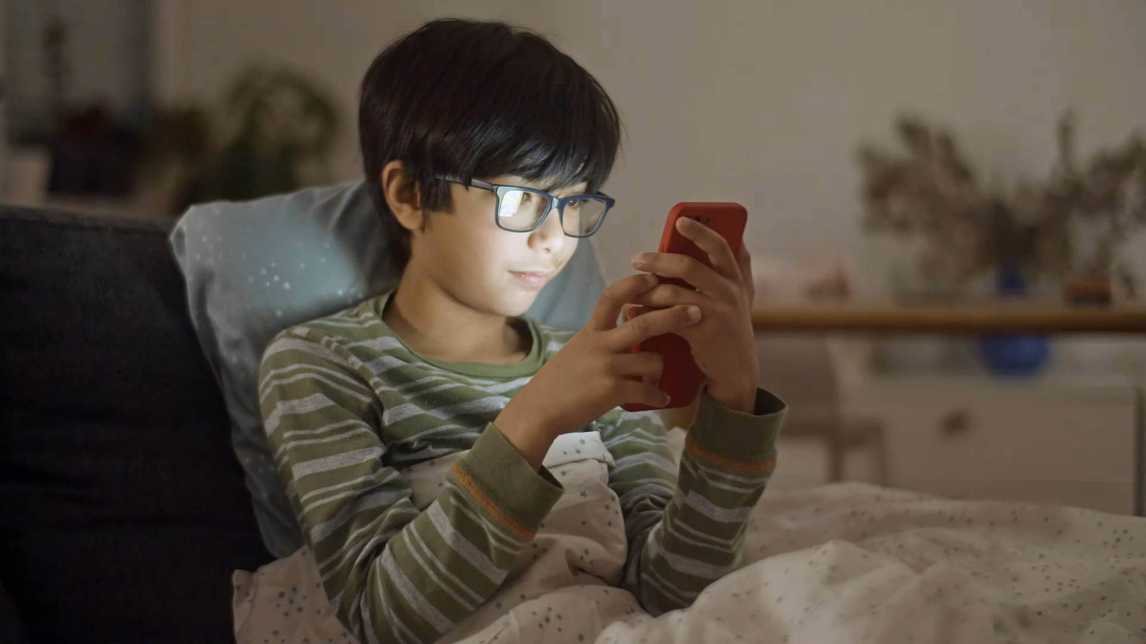 Child wearing glasses and striped pajamas looking at a red smartphone while sitting in bed with a blanket.