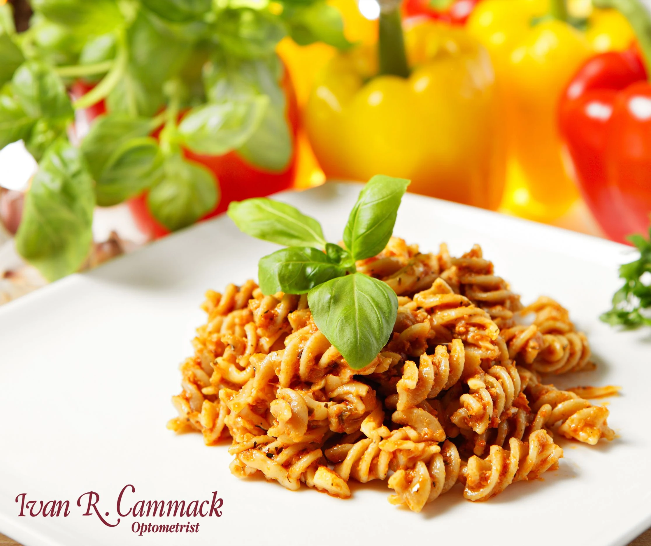 A serving of spiral pasta coated in red sauce is plated on a white dish and garnished with fresh basil leaves. Colorful bell peppers in red, yellow, and orange, along with fresh greens, are visible in the soft-focused background.