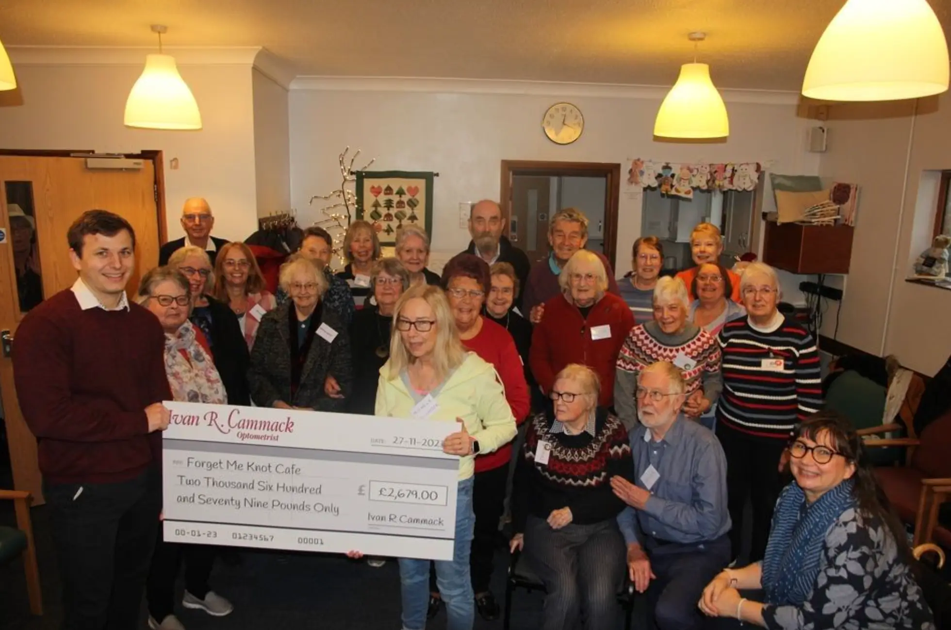 Group photo of people holding a large charity check for £2,679 made out to Forget Me Knot Cafe.