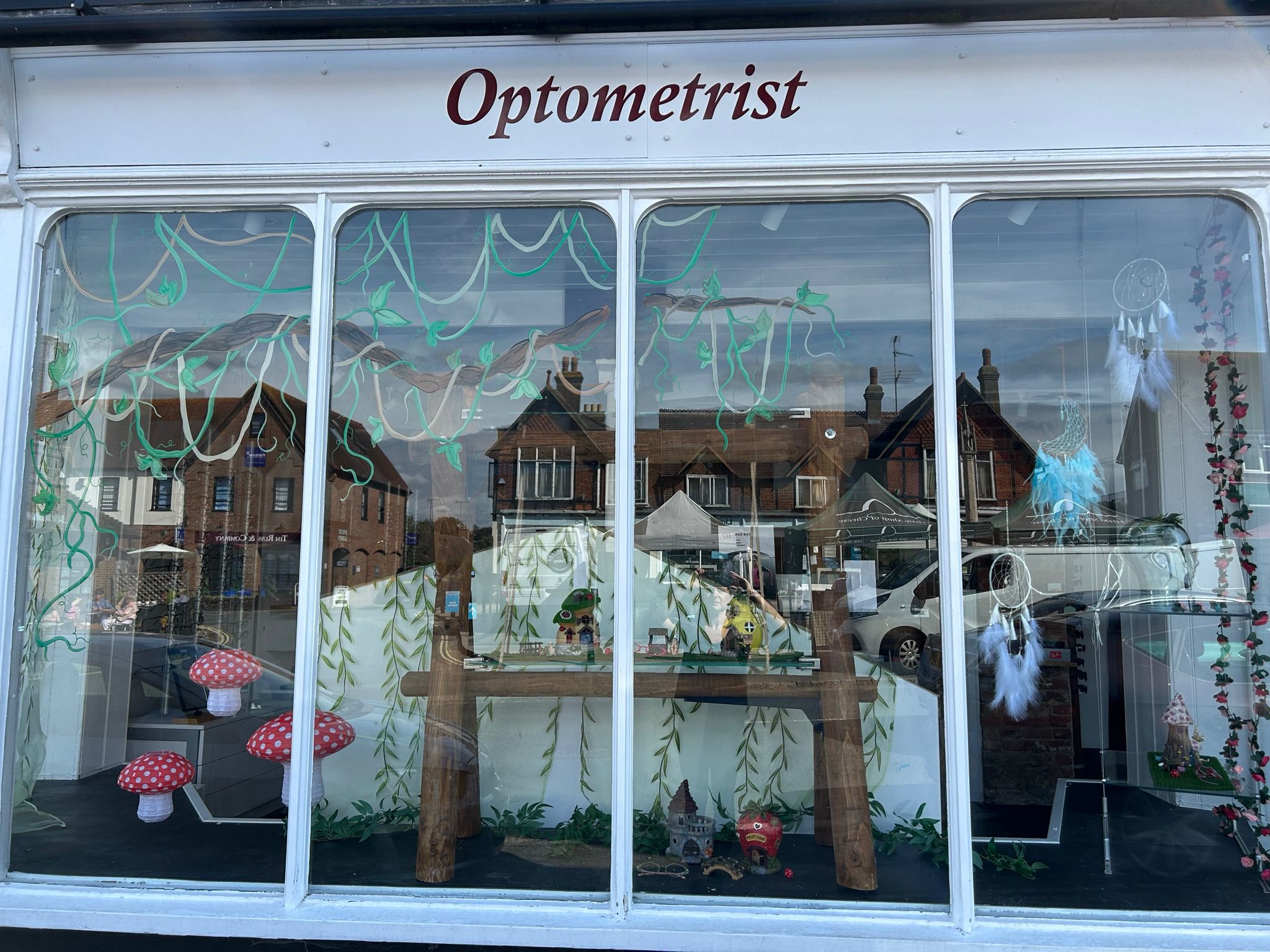 An enchanted forest-themed optometrist window display with painted willow branches, mushrooms, fairy houses, a wooden bridge structure, dreamcatcher, and whimsical decorative elements creating a magical woodland scene.