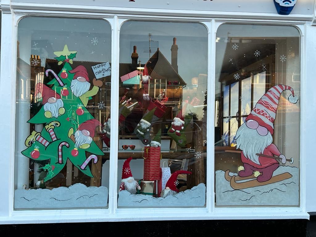 A holiday window display featuring playful gnome illustrations climbing on a Christmas tree, surrounded by presents and stockings, with another gnome sledding on the right panel.