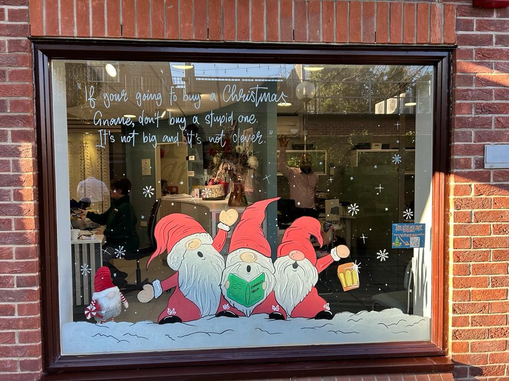 A humorous holiday window display featuring three cartoon gnomes in Santa outfits celebrating with drinks and a book, accompanied by text warning against buying stupid Christmas gnomes.