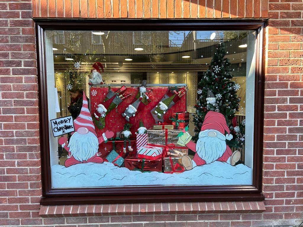 A festive storefront window display featuring two cartoon gnomes with red hats, wrapped presents, Christmas stockings, a decorated tree, and a "Merry Christmas" sign against a snowy scene.