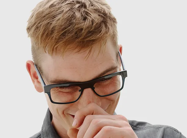 Young man wearing black-framed glasses looking down with a thoughtful expression against a white background.