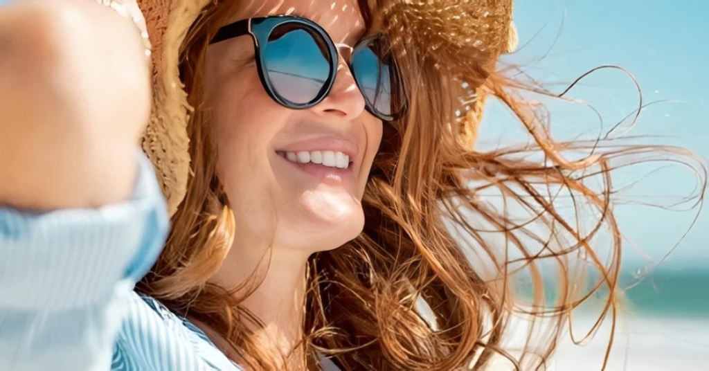 Woman wearing sunglasses and a straw hat smiling at the beach with wind-blown hair.
