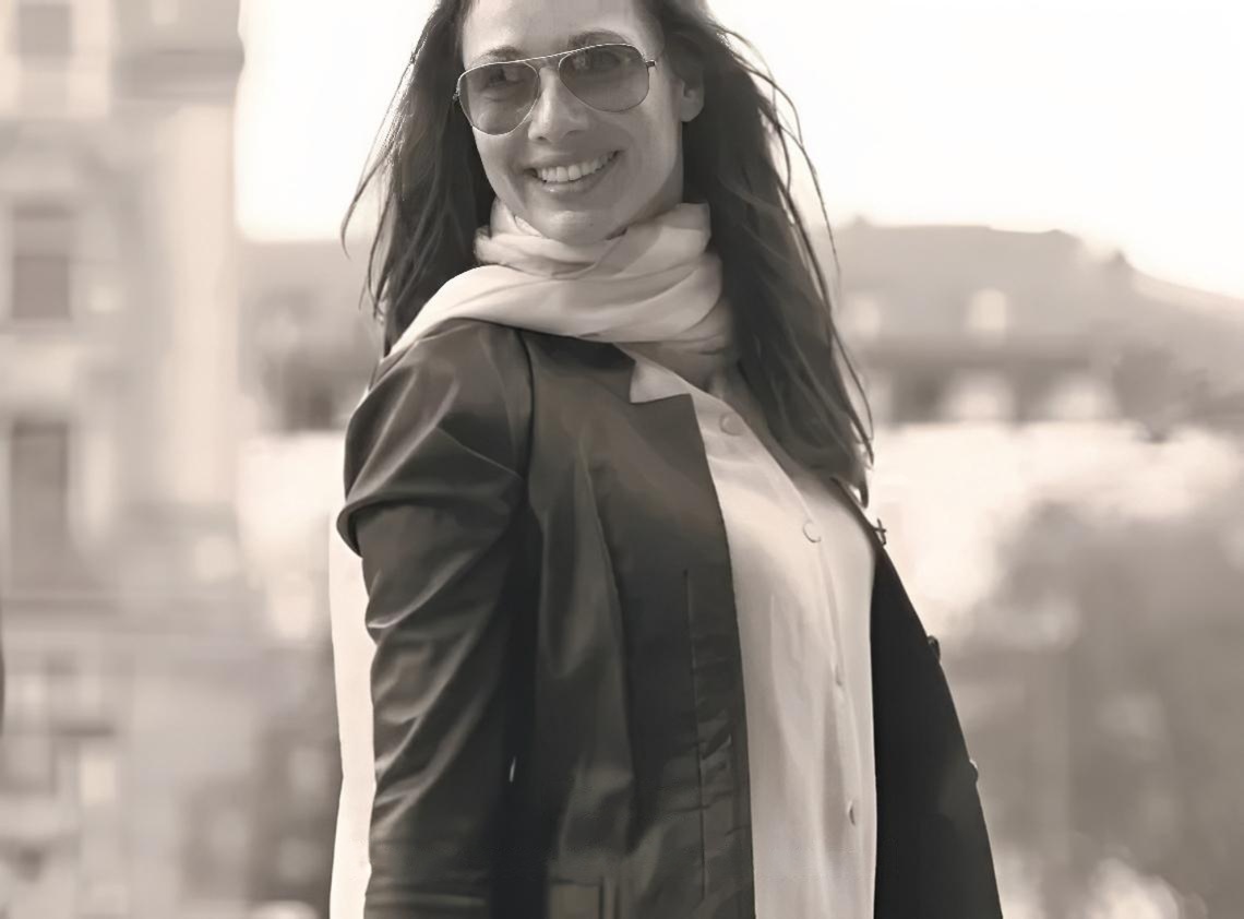 Black and white portrait of a smiling woman wearing aviator sunglasses and a scarf in an urban setting.