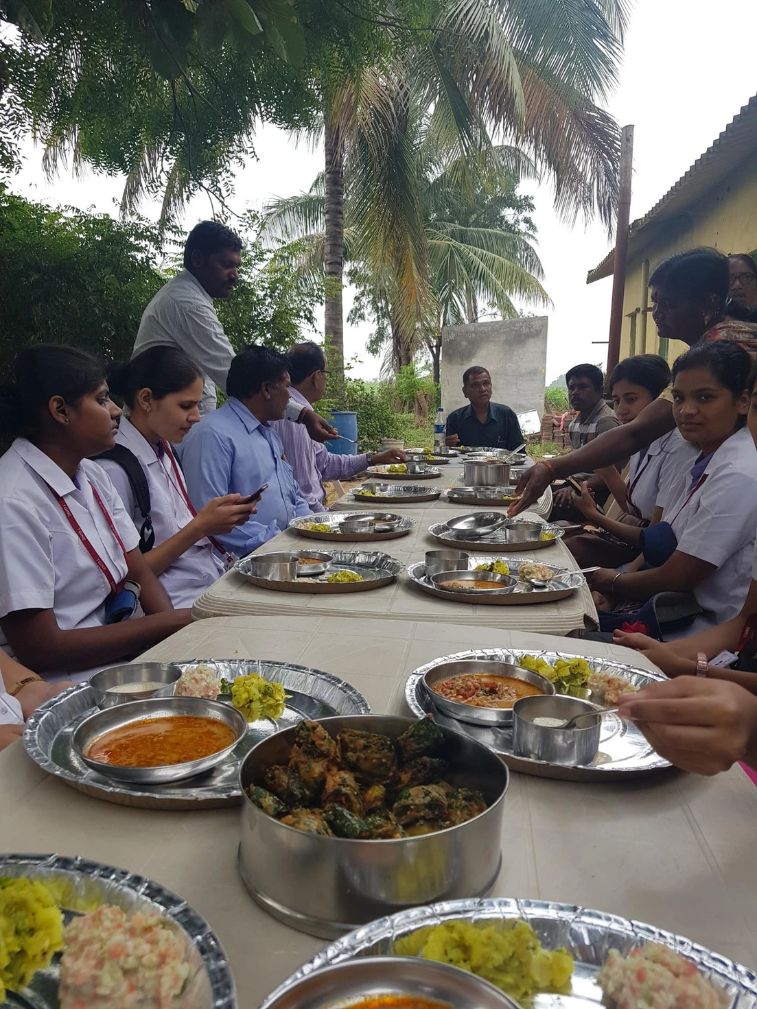 Students and community members gather outdoors under palm trees to share a communal meal served on metal plates at long tables.
