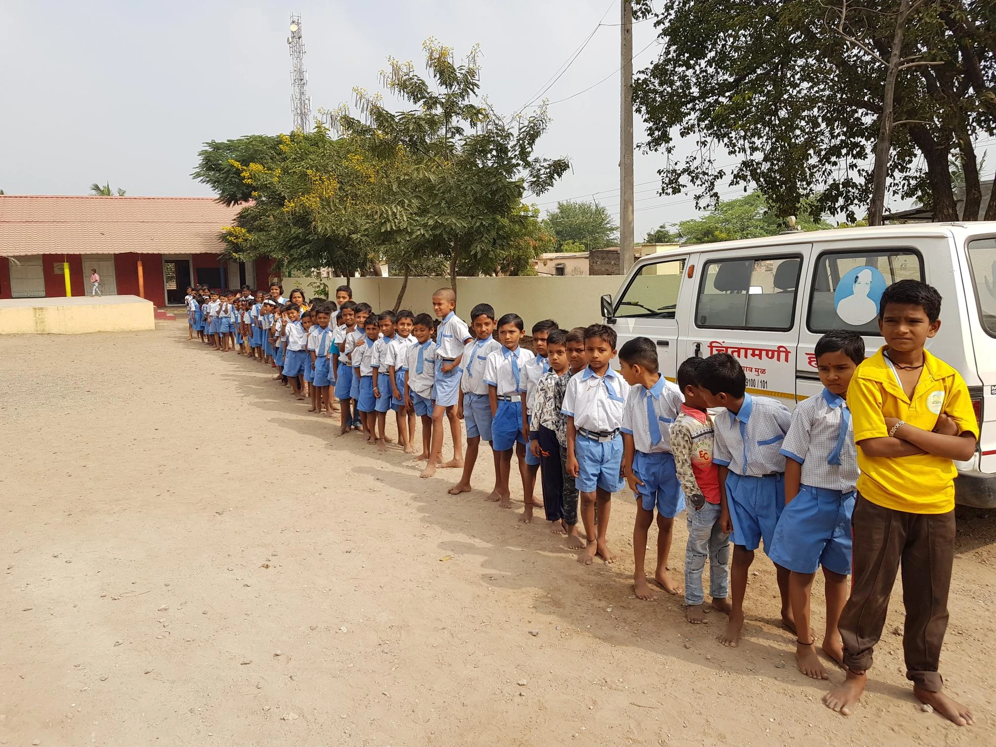 Students in school uniforms stand in a long queue beside a mobile health van at their rural school campus.