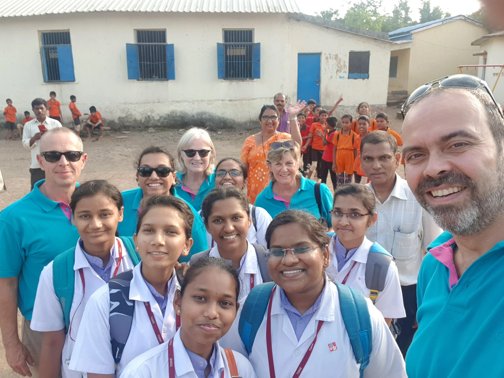 A group selfie of international volunteers in turquoise uniforms posing with smiling students and teachers at a rural school with children in orange shirts visible in the background.