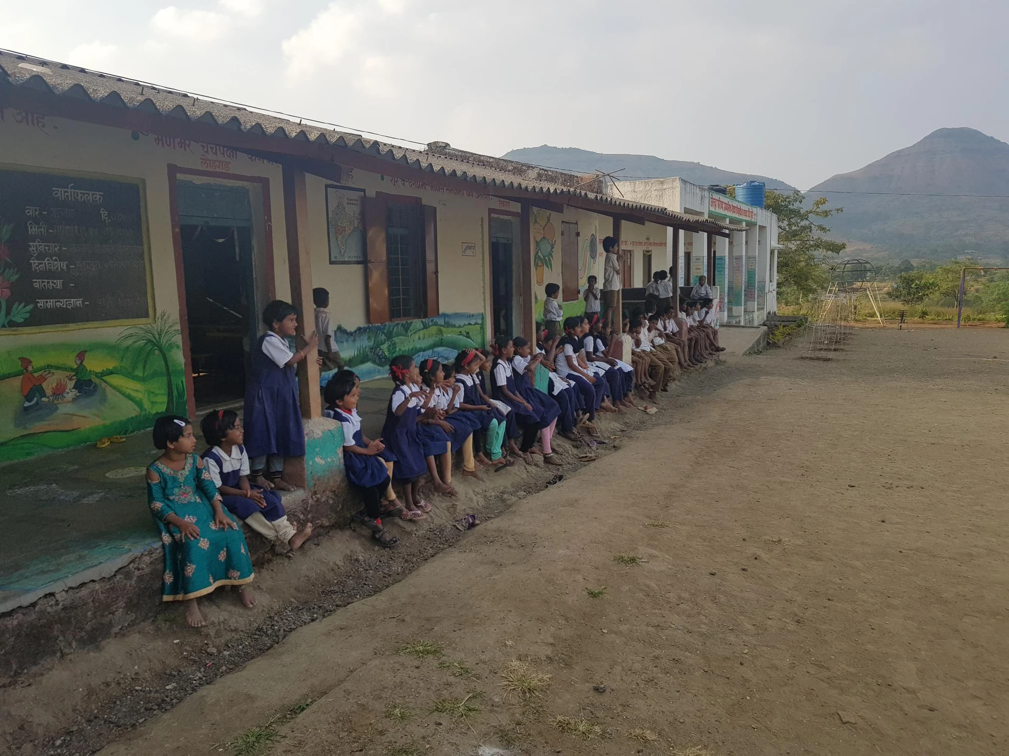 Students sit along the veranda of a rural school building decorated with colorful murals, with mountains visible in the background.