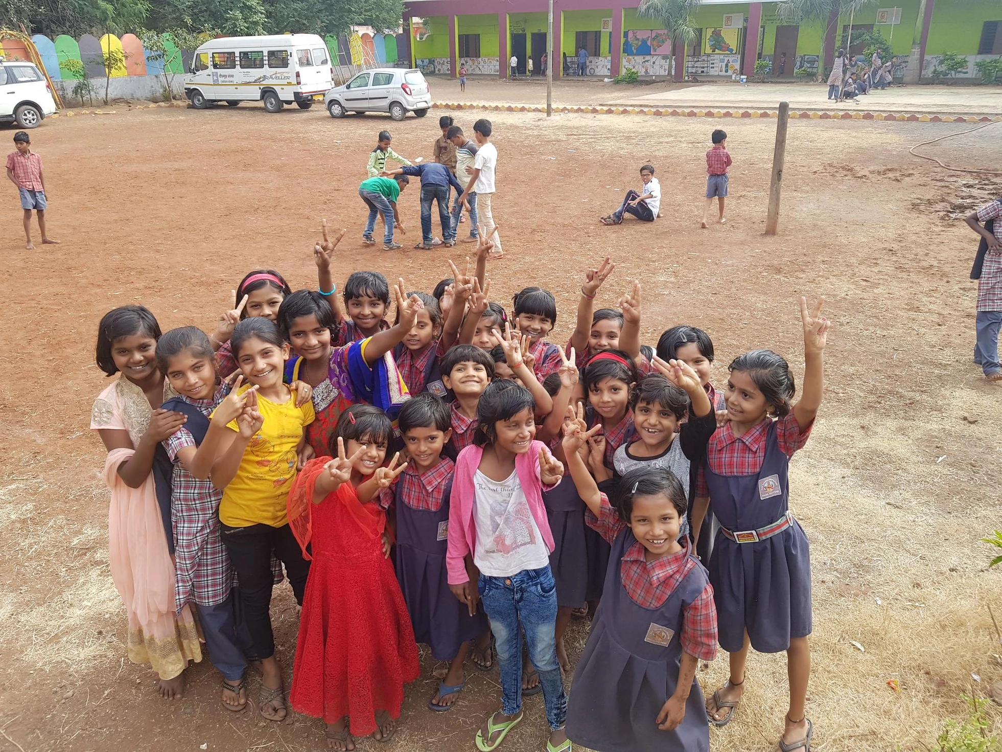 A cheerful group of schoolchildren pose with peace signs on a dirt playground in front of their colorful school building.