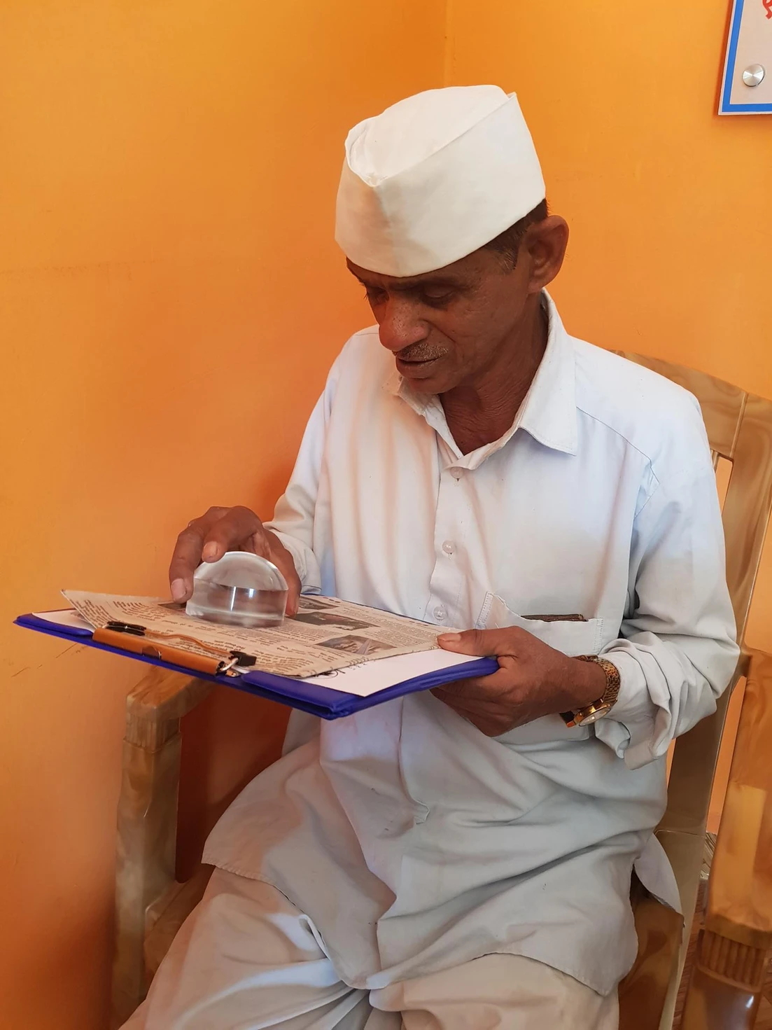 An elderly man in traditional white clothing and cap reads a newspaper using a magnifying glass while seated against an orange wall.