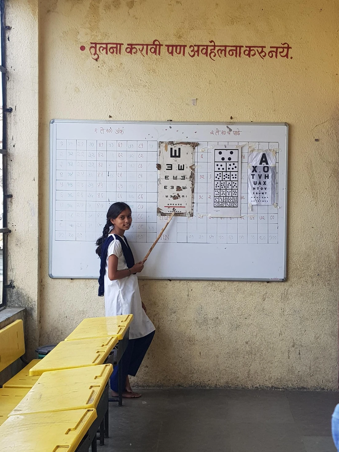 A young student in school uniform points to eye examination charts mounted on a whiteboard in a classroom with yellow benches.