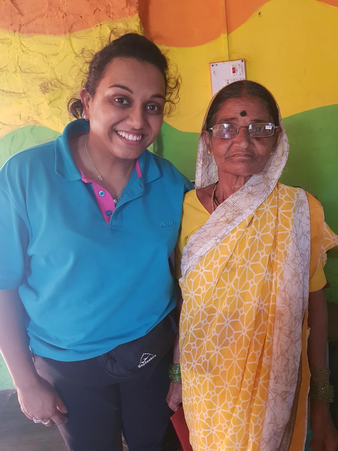 A young healthcare worker in a blue uniform poses with an elderly woman wearing glasses and a yellow traditional saree against a colorful painted wall.