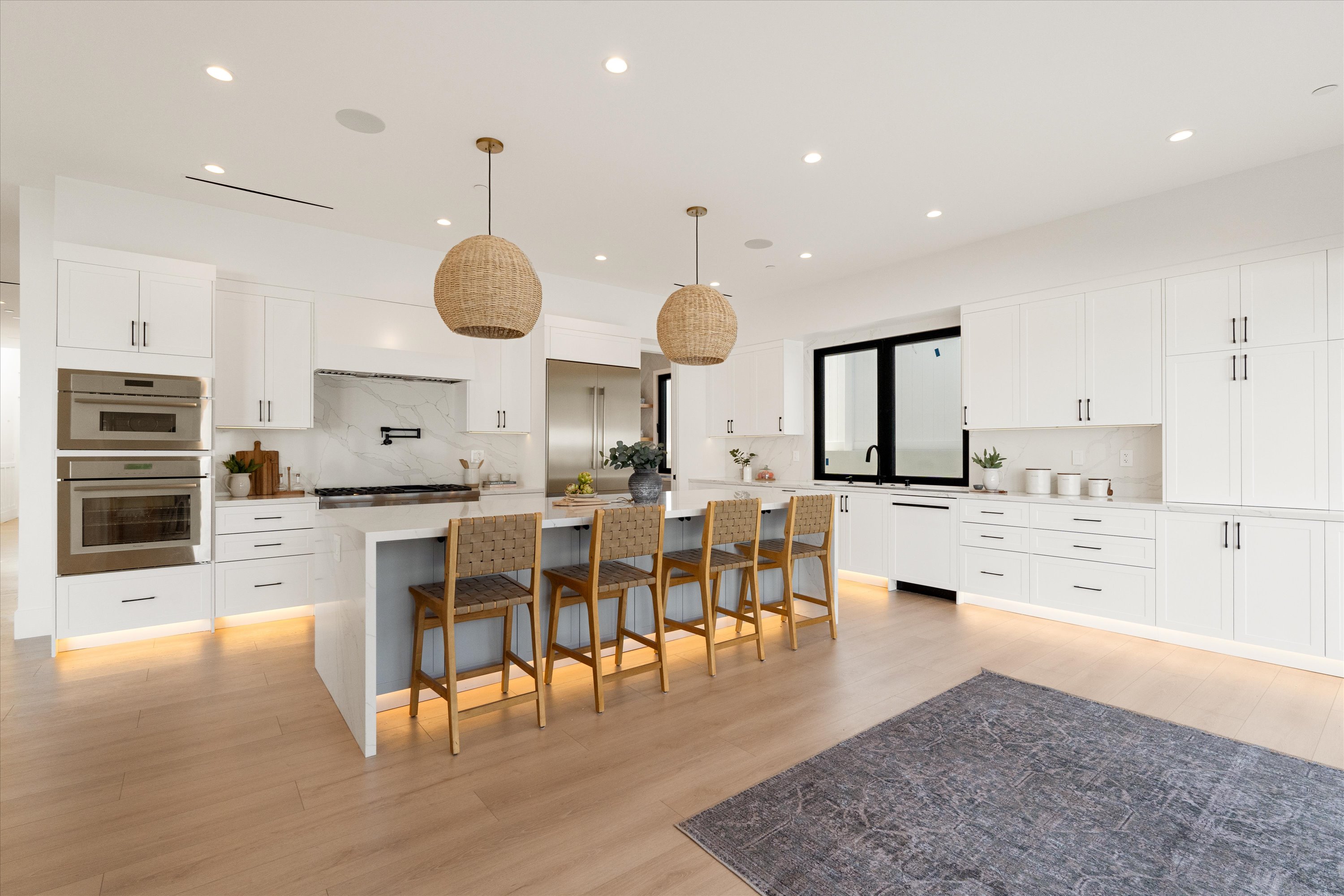Modern spacious kitchen with white cabinetry, a large island, woven counter chairs, pendant lights, and light hardwood floors.