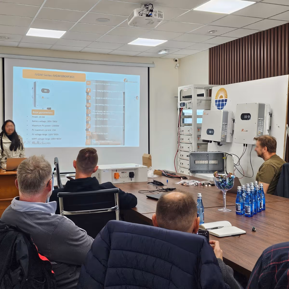 Szkolenie techniczne w siedzibie firmy Z-Ecoenergy. Prelegentka omawia specyfikację falownika serii IVGM wyświetlaną na ekranie, a uczestnicy siedzą przy stole naprzeciwko ściany z ekspozycją inwerterów i magazynów energii.