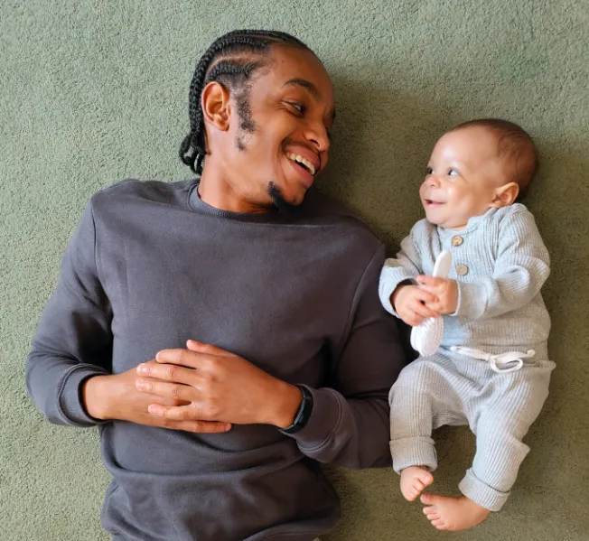 Smiling man with braided hair lying on carpet, looking at and holding hands with a baby in a gray outfit.