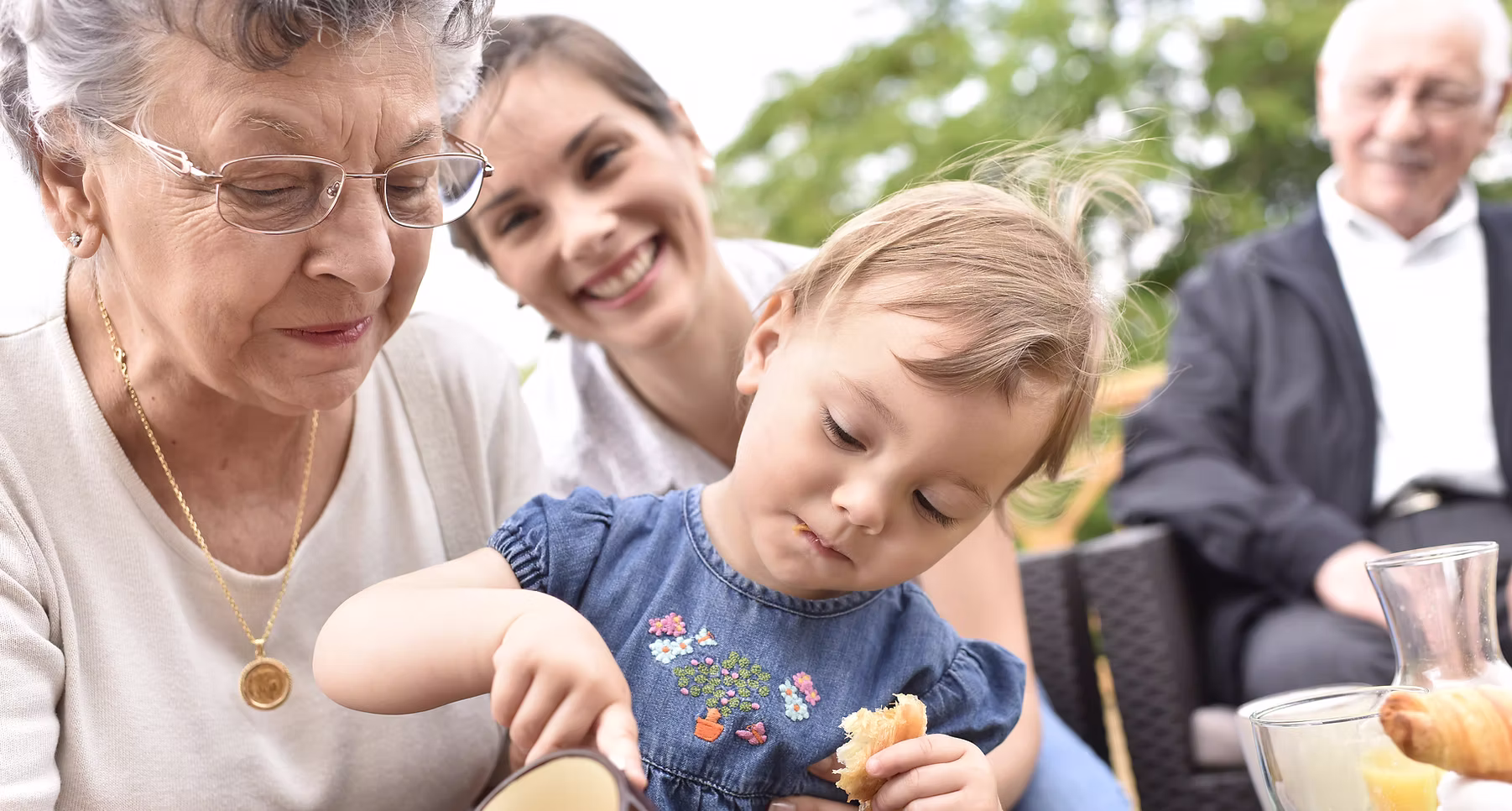A young child eating bread while sitting with a smiling woman and an older woman and man in the background outdoors.