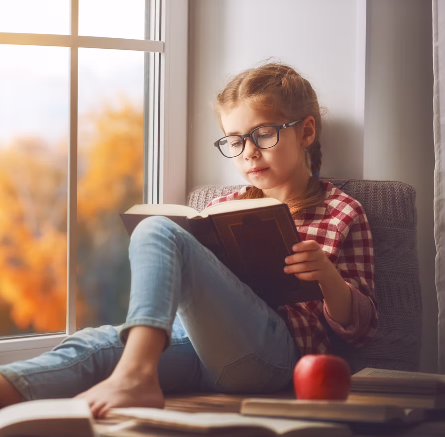 Young girl with glasses reading a book by a window with autumn trees outside.