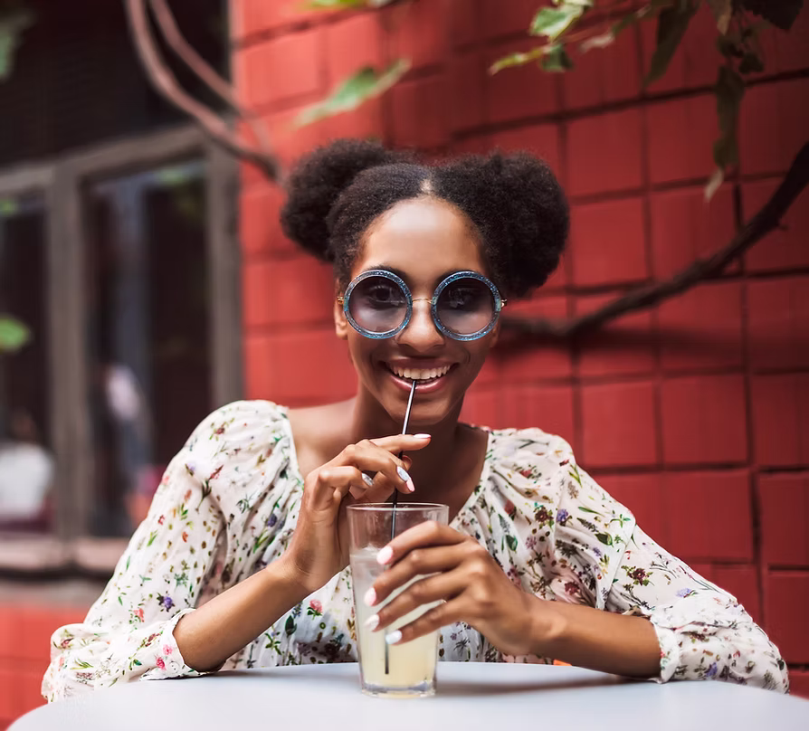 Young woman with afro puffs wearing round sunglasses and floral blouse drinking lemonade with a straw at an outdoor table.