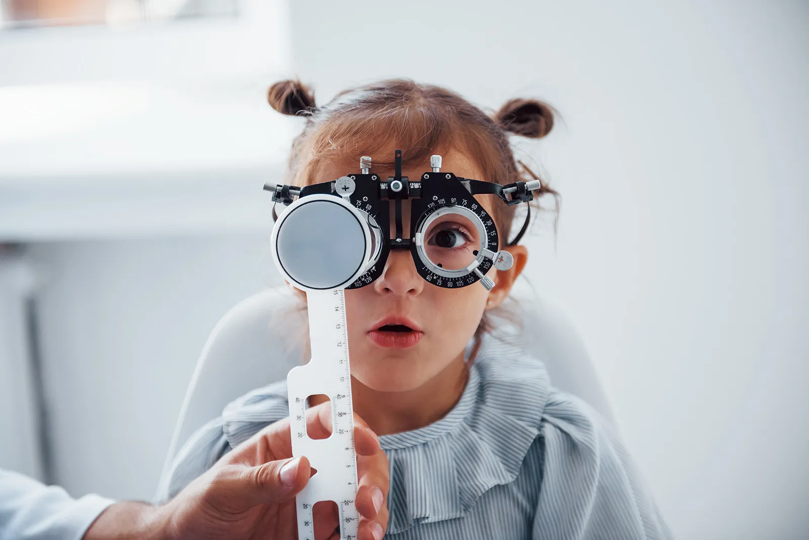 Young girl with hair buns having her eyes tested with a phoropter and measuring ruler during a children's eye examination