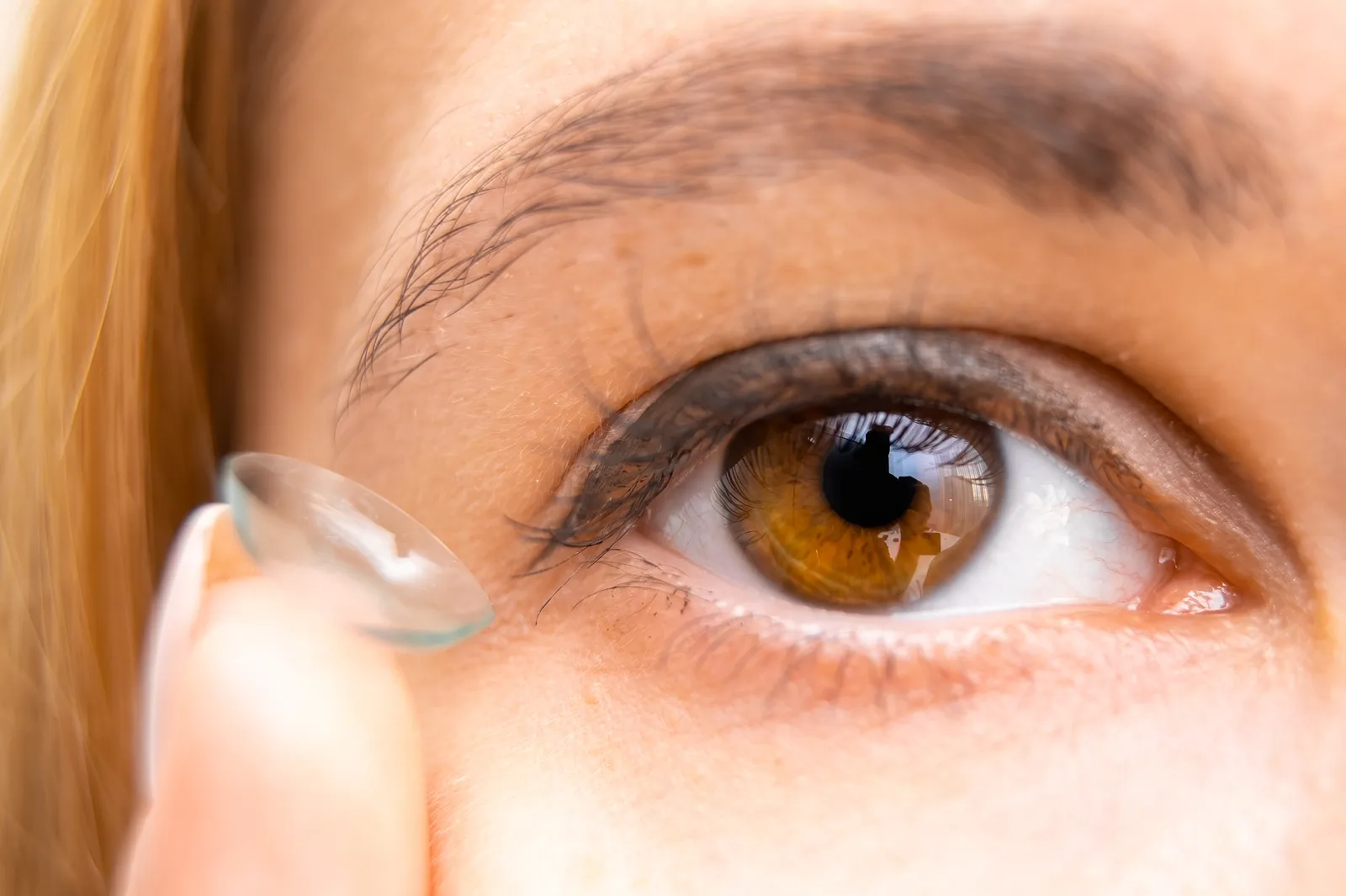 Close-up of a woman's brown eye as she holds a contact lens on her fingertip, ready to apply it