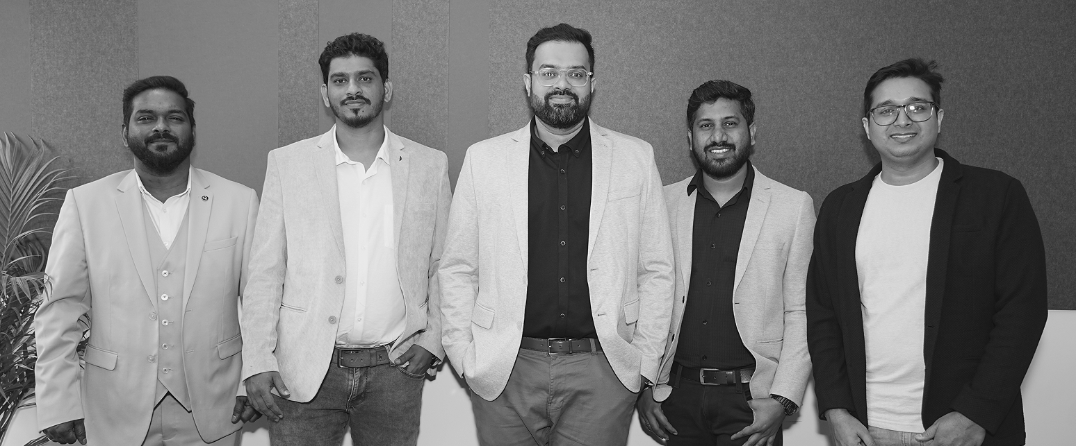 Five men standing side by side wearing blazers and smiling in front of a wall with paneling.