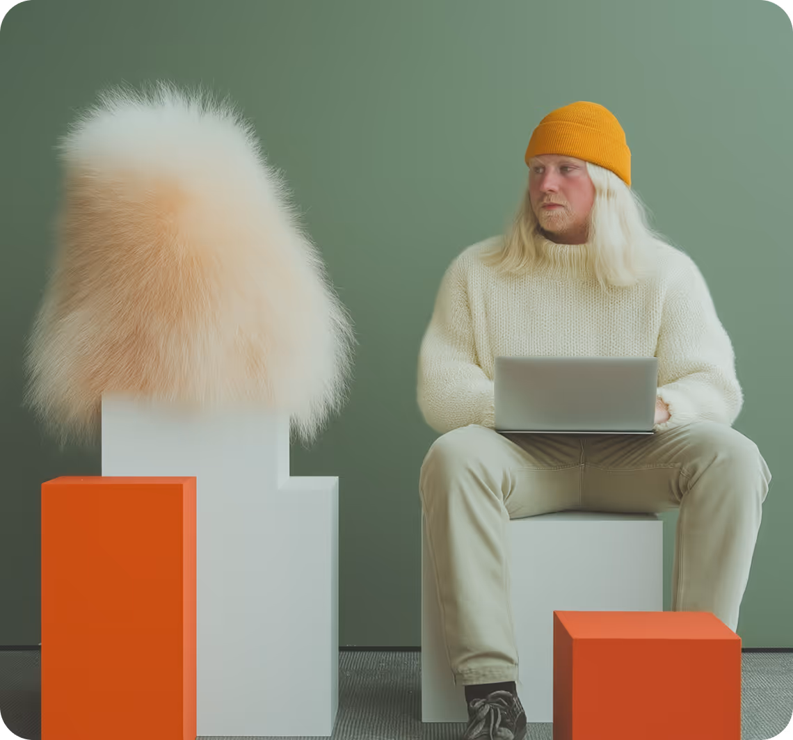 Man in a yellow beanie and cream sweater sitting on a white block with a laptop, looking to the side next to a furry white and beige object on a white pedestal and two orange cubes.