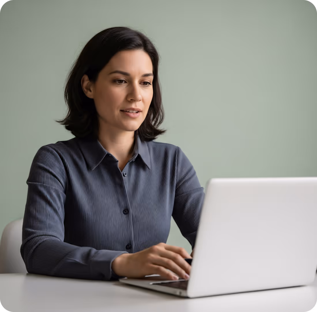 Woman with dark hair wearing a gray button-up shirt working on a silver laptop at a white desk.