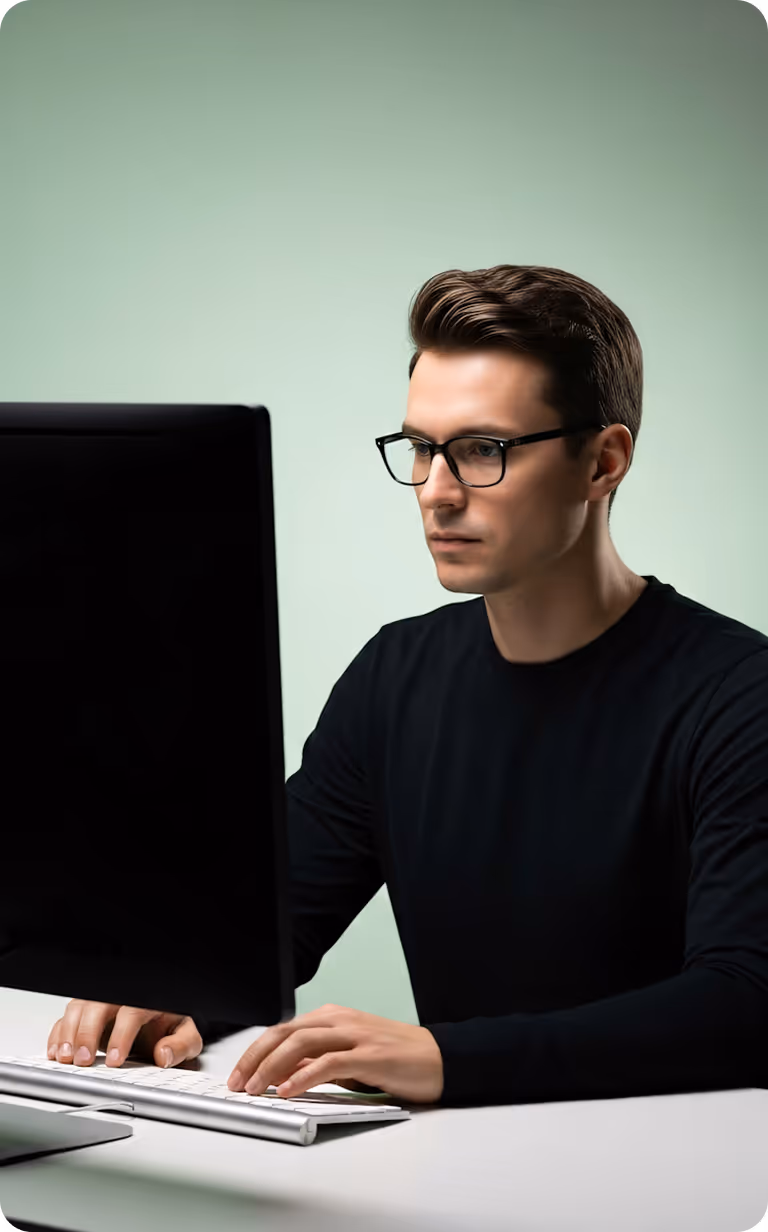 Man with glasses focused on working at a desktop computer with a wireless keyboard on a white desk.