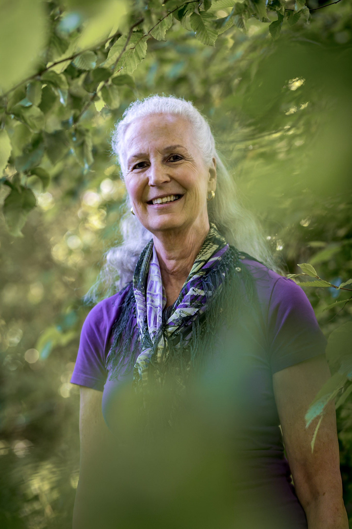 Smiling elderly woman with long white hair, wearing a purple shirt and patterned scarf, surrounded by green foliage.