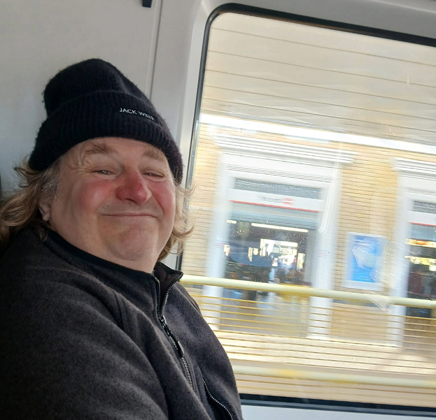 Smiling man wearing a black beanie and dark jacket sitting inside a vehicle next to a window showing a blurred cityscape outside.