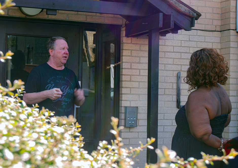 A man in a black t-shirt speaking to a woman with curly hair wearing a black strapless dress outside a brick building entrance.
