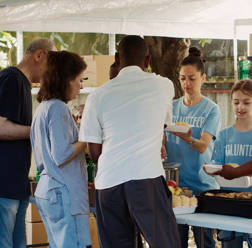 Volunteers in blue shirts serving food to people at an outdoor food distribution stand.