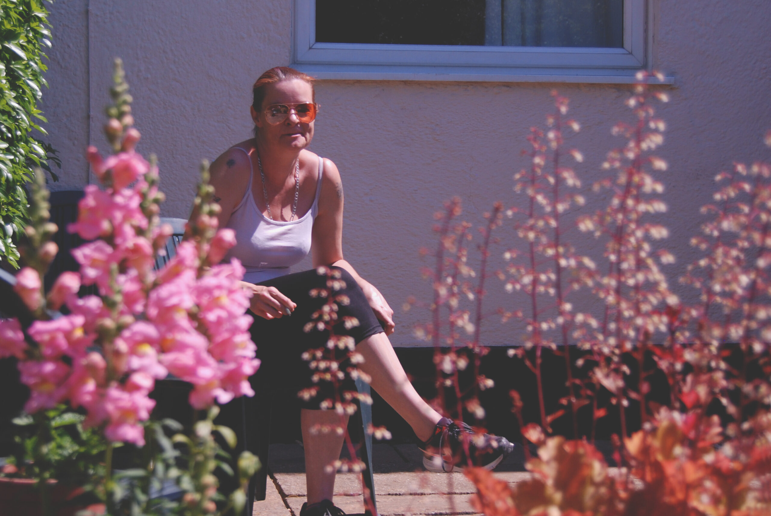 Woman wearing sunglasses sitting outside on a chair near a wall and window, with pink flowers in the foreground.